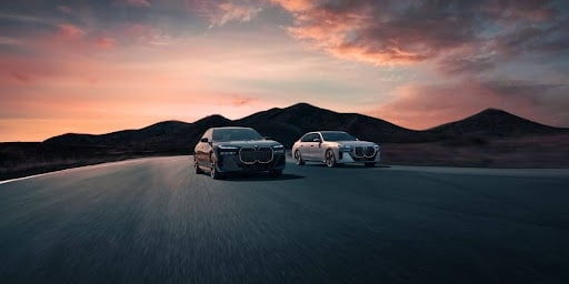 Two BMWs with rear cross traffic alert driving down a highway at sunset.