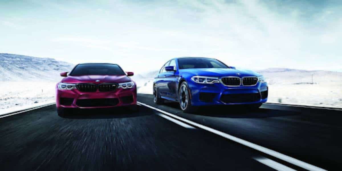 Red and blue 2025 bmw m5 driving next to eachother down a highway with a snowy backdrop.