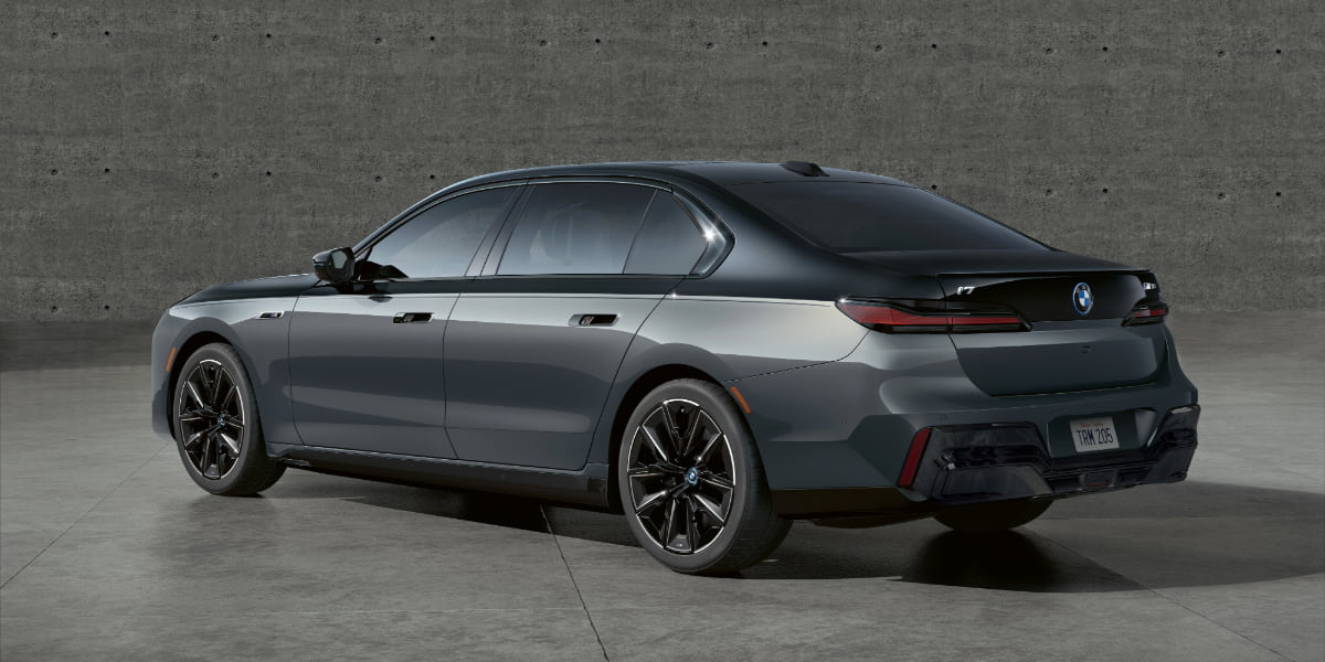 A rear three-quarter view of the 2025 BMW i7, highlighting its sculpted body, LED taillights, and aerodynamic design against a modern concrete backdrop.