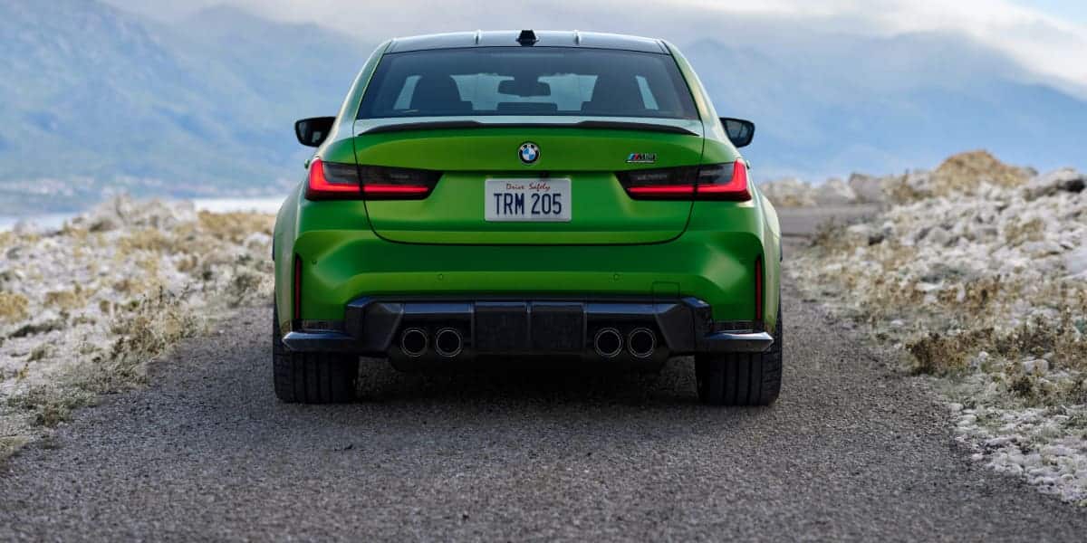 A rear view of the 2025 BMW M3 parked on a rugged landscape, emphasizing its wide stance, LED taillights, and signature quad exhaust system.