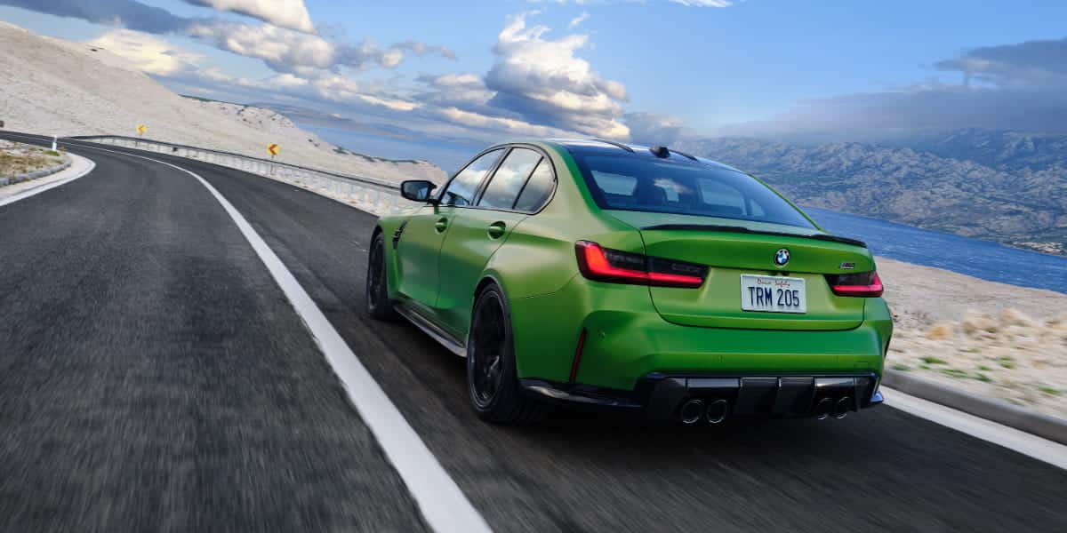 The 2025 BMW M3 in motion on a winding coastal road, showcasing its aggressive rear design, quad exhaust tips, and dynamic performance.