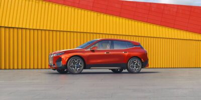 Side view of a red BMW iX parked in front of a bold yellow and red building, highlighting the design and size of the iX electric SUV.