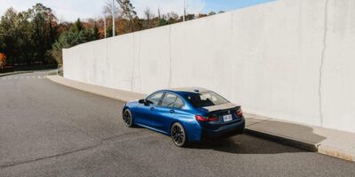 Rear three quarter view of a blue 2026 BMW 3 series sport sedan driving on a city street beside a tall concrete wall